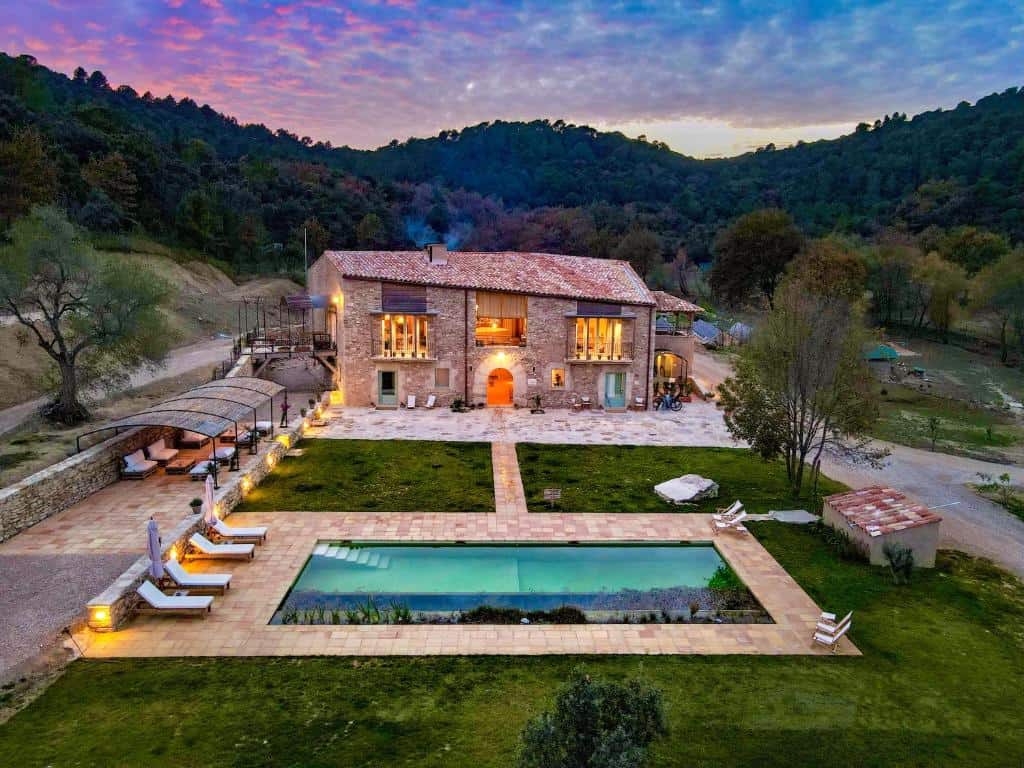 Villa de piedra con ventanas iluminadas, rodeada de árboles, cuenta con una piscina rectangular, tumbonas y una zona de patio al atardecer.