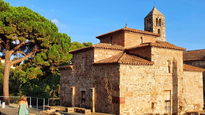 Una mujer se sienta en un banco cerca de una histórica iglesia de piedra con campanario, rodeada de árboles bajo un cielo azul despejado.